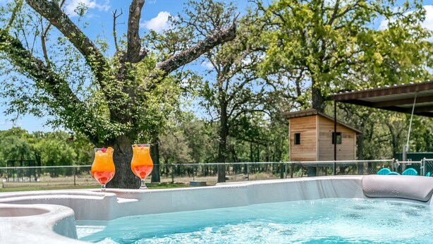 A swimming pool with two glasses of orange drink, surrounded by trees and a wooden cabin.