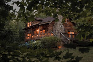 A log cabin with a stone foundation, wooden exterior, and a porch with lights.