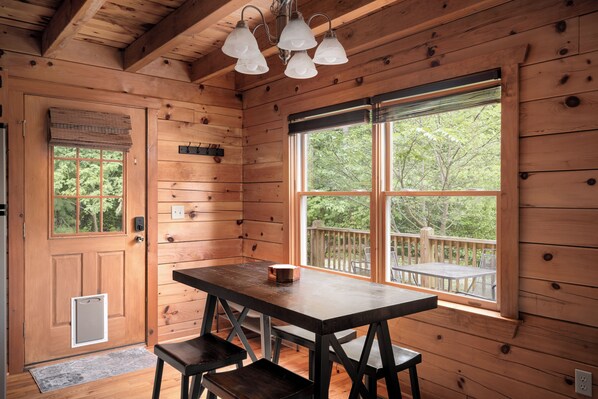 A wooden cabin interior with a dining table, benches, a window with a view of trees, and a door with a mailbox.