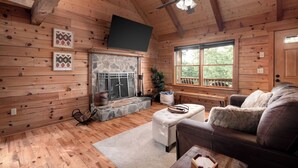 A wooden cabin interior with a stone fireplace, a ceiling fan, and a large window. - Shenandoah Log Cabin *Hot Tub* *Indoor Fireplace* (Dyke)