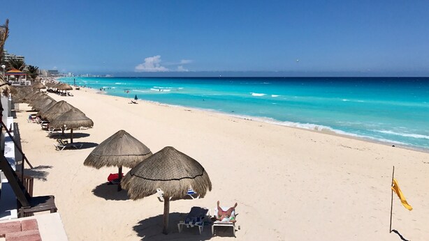 A beach with thatched umbrellas and lounge chairs, a clear blue sea, and a distant city skyline. - Brisas Beachfront Penthouses (Cancun)