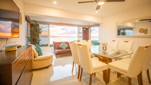 A modern living room with a glass dining table, white chairs, a sofa, and a large window with a view of the sunset. - Brisas Beachfront Penthouses (Cancun)
