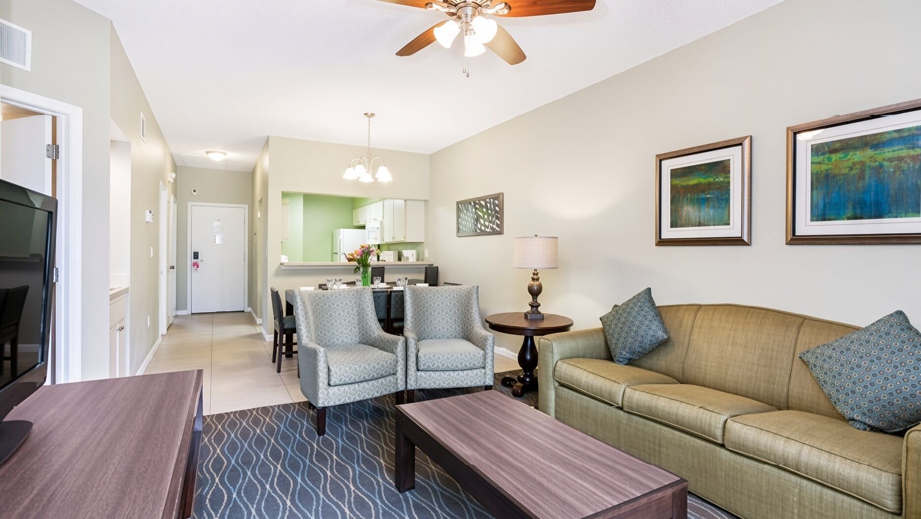 A living room with a sofa, armchairs, a coffee table, a dining area with chairs, a kitchen in the background, and a ceiling fan.