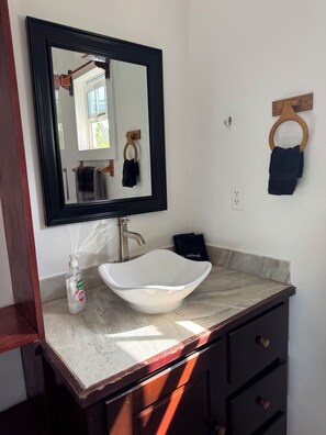 A bathroom with a white vessel sink, a large mirror, a wooden towel rack, and a dark-colored towel. - Secret Beach Cabanas, Green Cabana. Sea Views just steps to water access! (San Pedro)