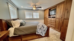 Een slaapkamer met een bed, een houten kledingkast, plafondventilator en twee ramen met jaloezieën.