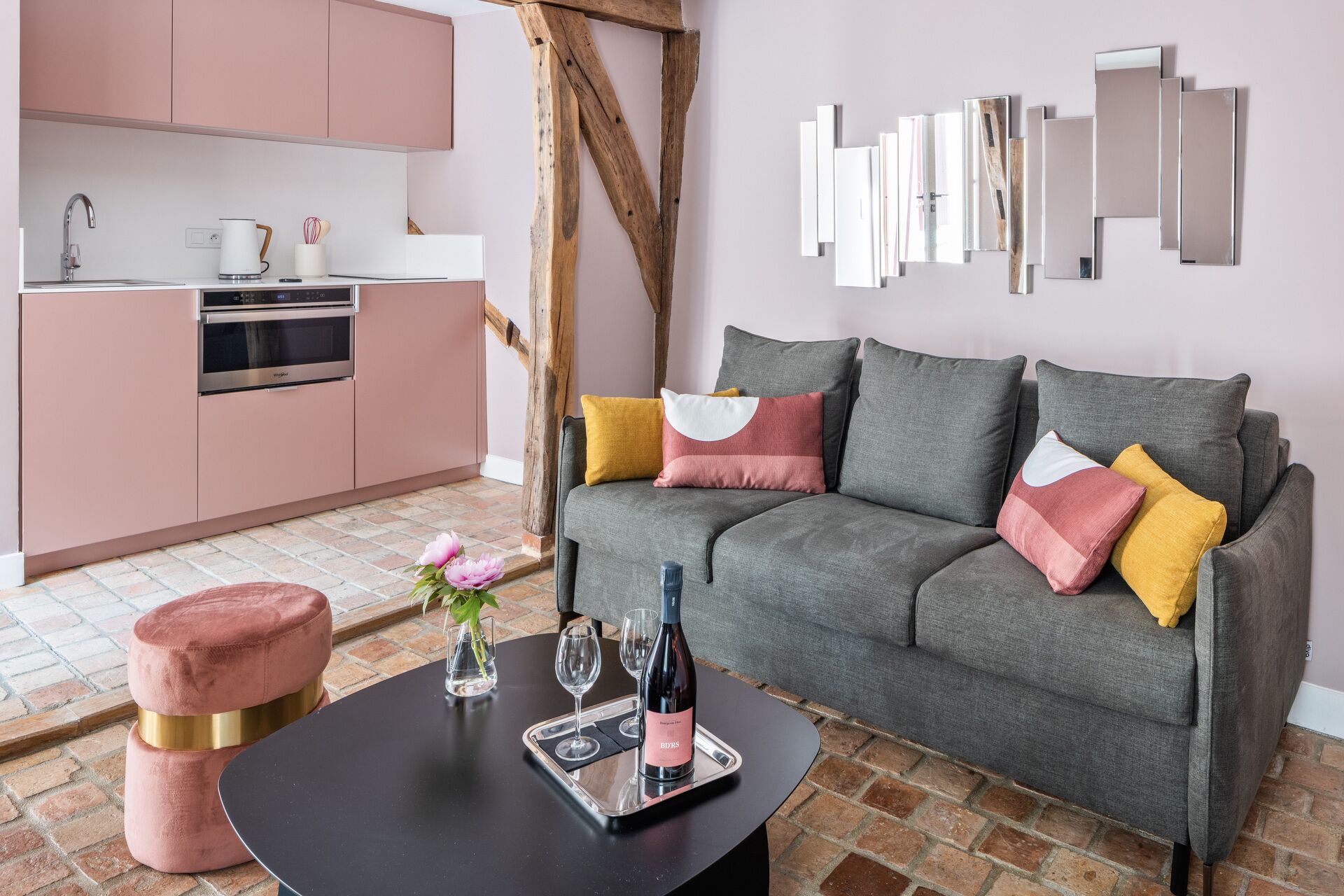 A modern living room with a grey sofa, pink and yellow cushions, a black coffee table, and a kitchen area with pink cabinets and a wooden beam.