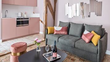 A modern living room with a grey sofa, pink and yellow cushions, a black coffee table, and a kitchen area with pink cabinets and a wooden beam.
