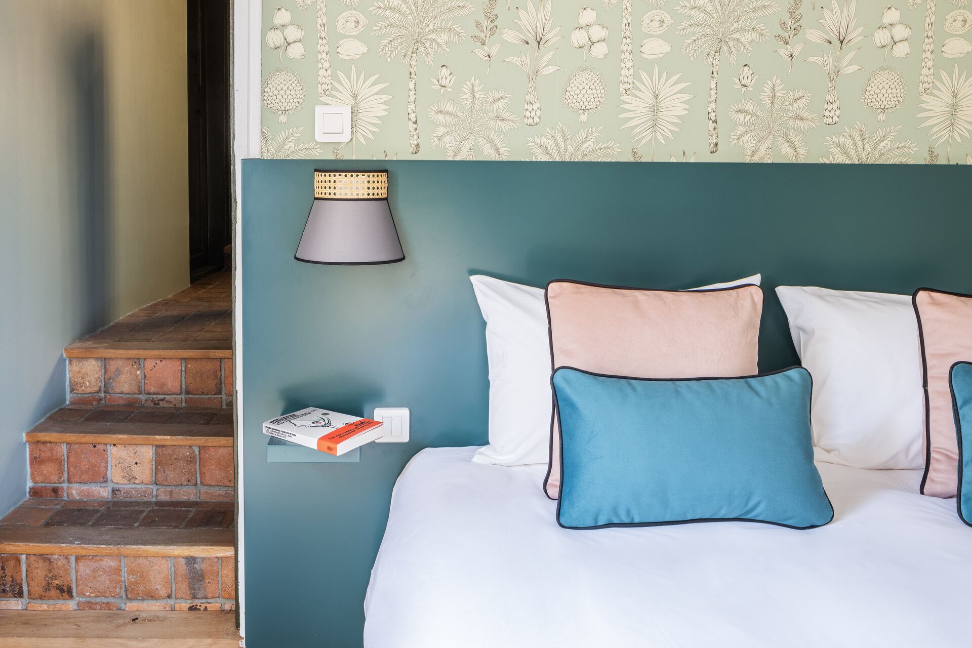 A bedroom with a bed, pillows, a nightstand, a lamp, and a wall with a botanical pattern.