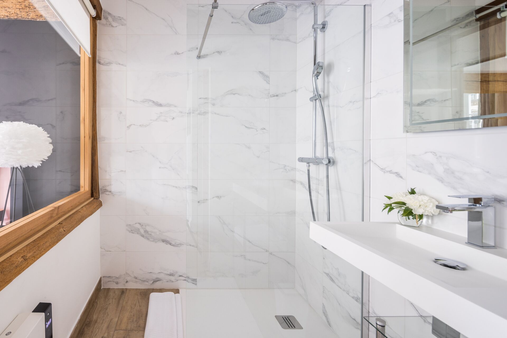 A modern bathroom with a shower, marble walls, a wooden floor, and a white sink with a mirror.