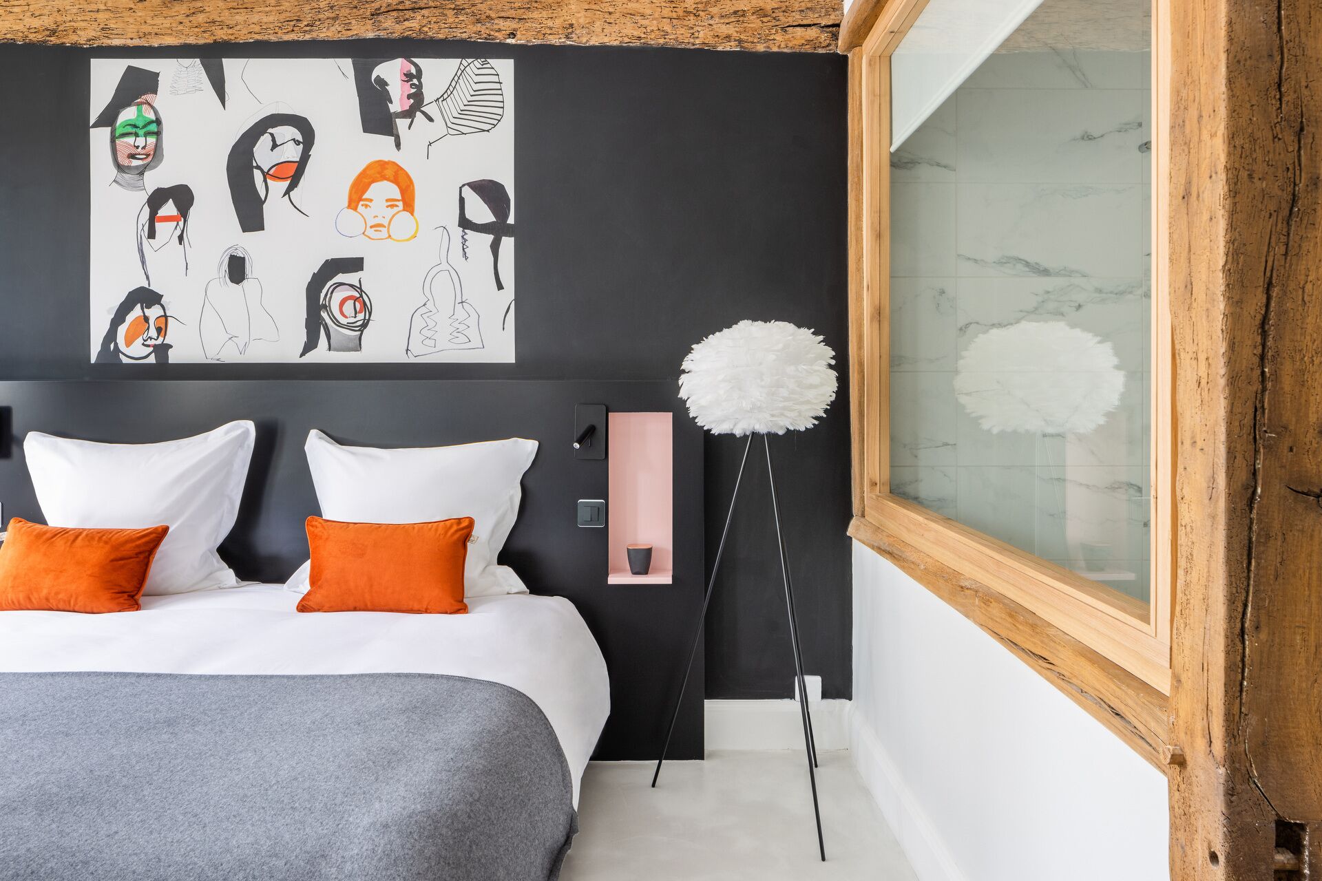 A modern bedroom with a large bed, two orange pillows, a gray throw, a black wall with a white abstract art piece, a tall white floor lamp, and a wooden-framed window.