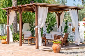 Una pérgola de madera con cortinas blancas, un banco acolchado y una silla de madera.