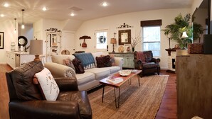 Ein gemütliches Wohnzimmer mit Ledersofa, weißem Couch und Holztisch. Es gibt eine Küchenzeile mit Kühlschrank, Spüle und Arbeitsplatte. An der Wand hängt ein großer Spiegel, und im Eck steht ein Blumentopf.