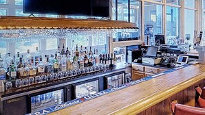 Eine Bar mit einer Holztresen, Regalen voller Spirituosenflaschen, einem Fernseher über der Bar und Blick durch große Fenster nach draußen.