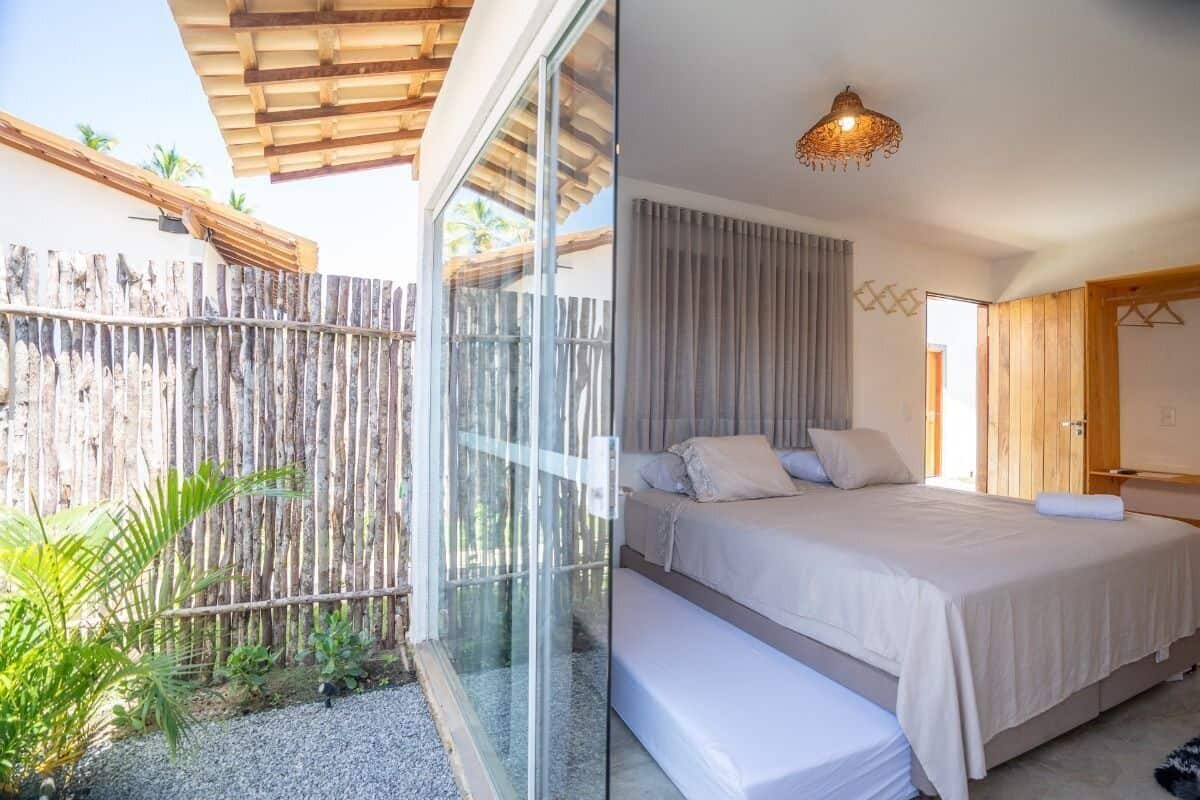 A bedroom with a large bed, a wooden ceiling, and a glass door leading to a balcony with a bamboo fence and plants.