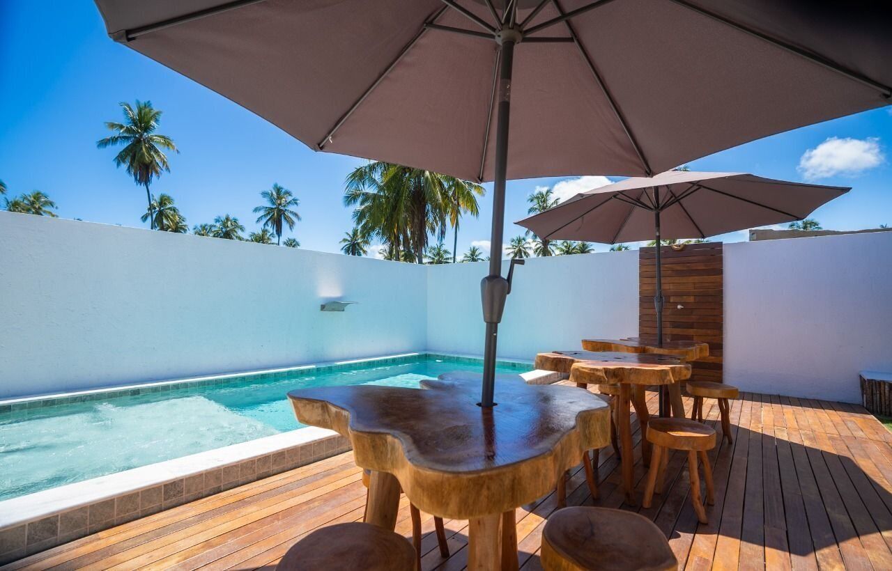 Un espace aménagé au bord de la piscine, avec des tables et des chaises en bois, un grand parasol et une vue sur les palmiers.