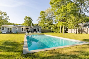Una piscina rettangolare con superficie d'acqua cristallina e blu, circondata da un prato curato e da una casa con tetto spiovente e un camino.