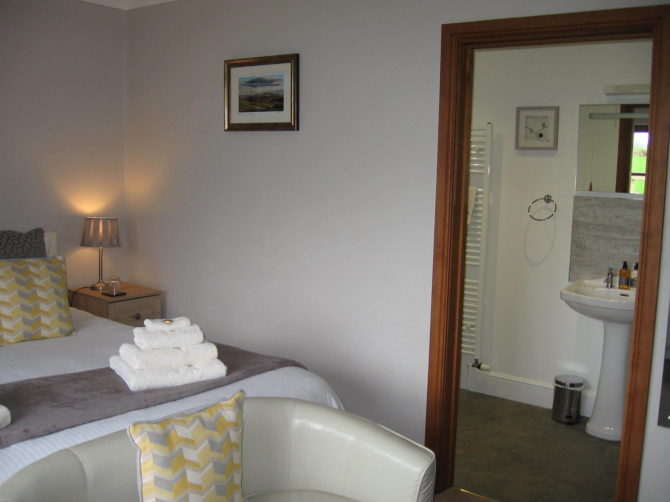 Habitación de hotel con cama, mesita de noche con lámpara, una silla, baño visible a través de una puerta abierta y un cuadro en la pared.