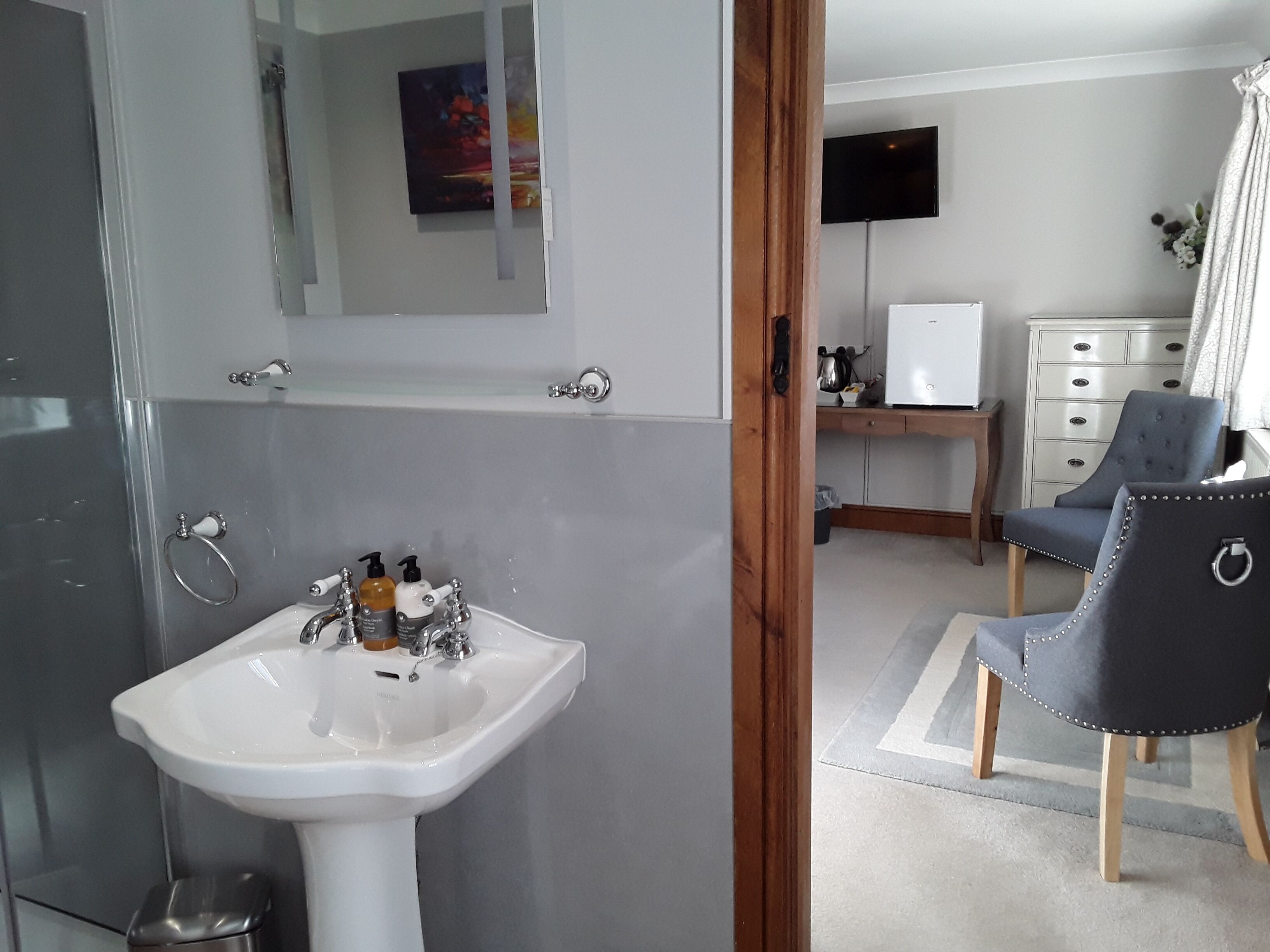 Een badkamer met een wit wastafelmeubel, een spiegel en een douche. Er is een televisie aan de muur en een stoel in de kamer.