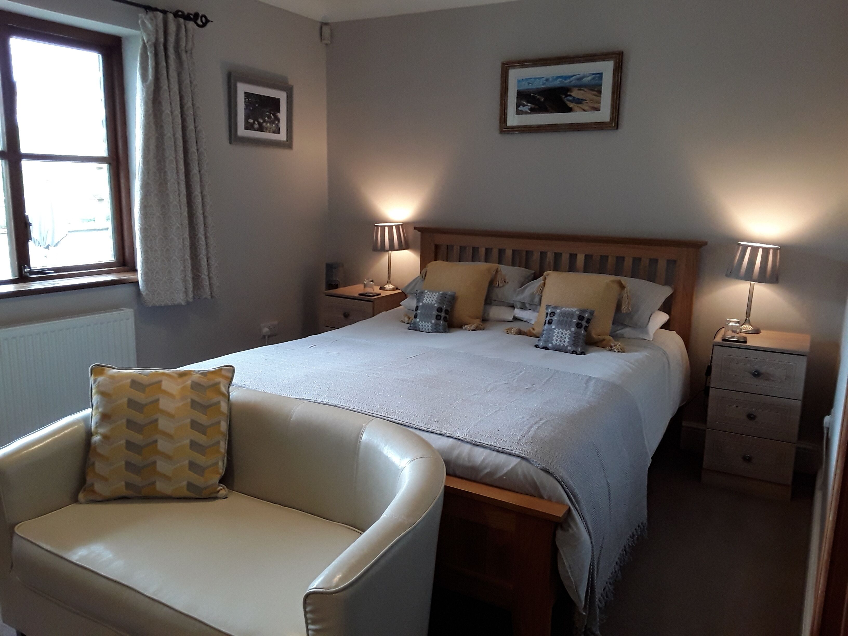 A bedroom with a bed, two bedside tables with lamps, a chair, a window with curtains, and two framed pictures on the wall.