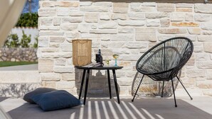A stone wall with a wooden panel above, a round table with a bottle and glass, a black woven chair, and a blue cushion on the ground. - Villa Luna with heated swimming pool & E-bikes (Županje (Krk))