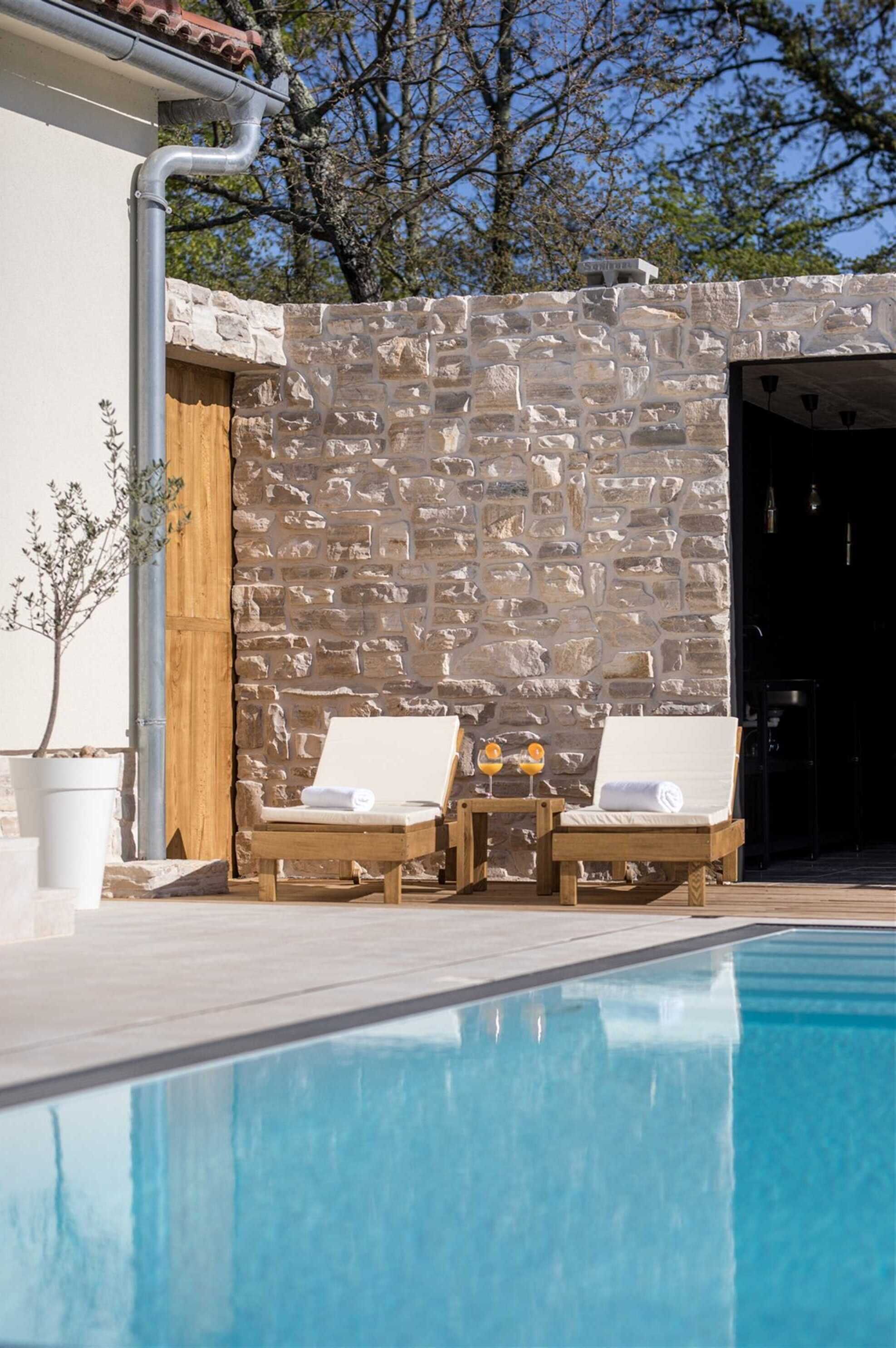 A pool area with two lounge chairs, a stone wall, and a potted plant.