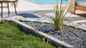 A swimming pool with a stone structure in the background, a grassy area with plants, and a seating area with a chair and table. - Villa Luna with heated swimming pool & E-bikes (Županje (Krk))