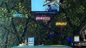 Een hotel receptie met een receptiebalie van bamboe, een televisiescherm en een wand versierd met kunstplanten en neonborden.