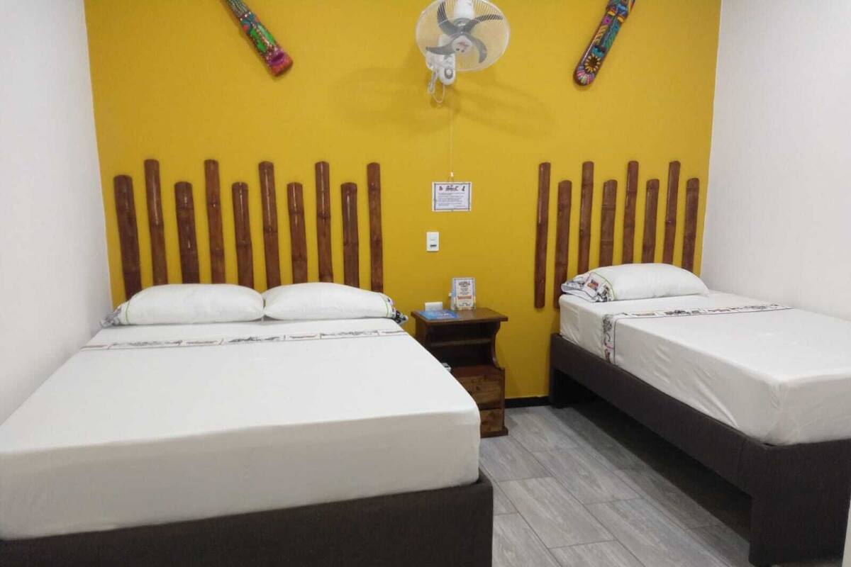 Ein Zimmer mit zwei Betten, einem kleinen Tisch, einem Ventilator und einer Wand mit vertikalen Holzlatten.