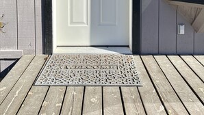 En vit dörr med ett litet fönster ovanför, en dörrmatta med ordet ”VÄLKOMMEN” och en lampa på väggen.