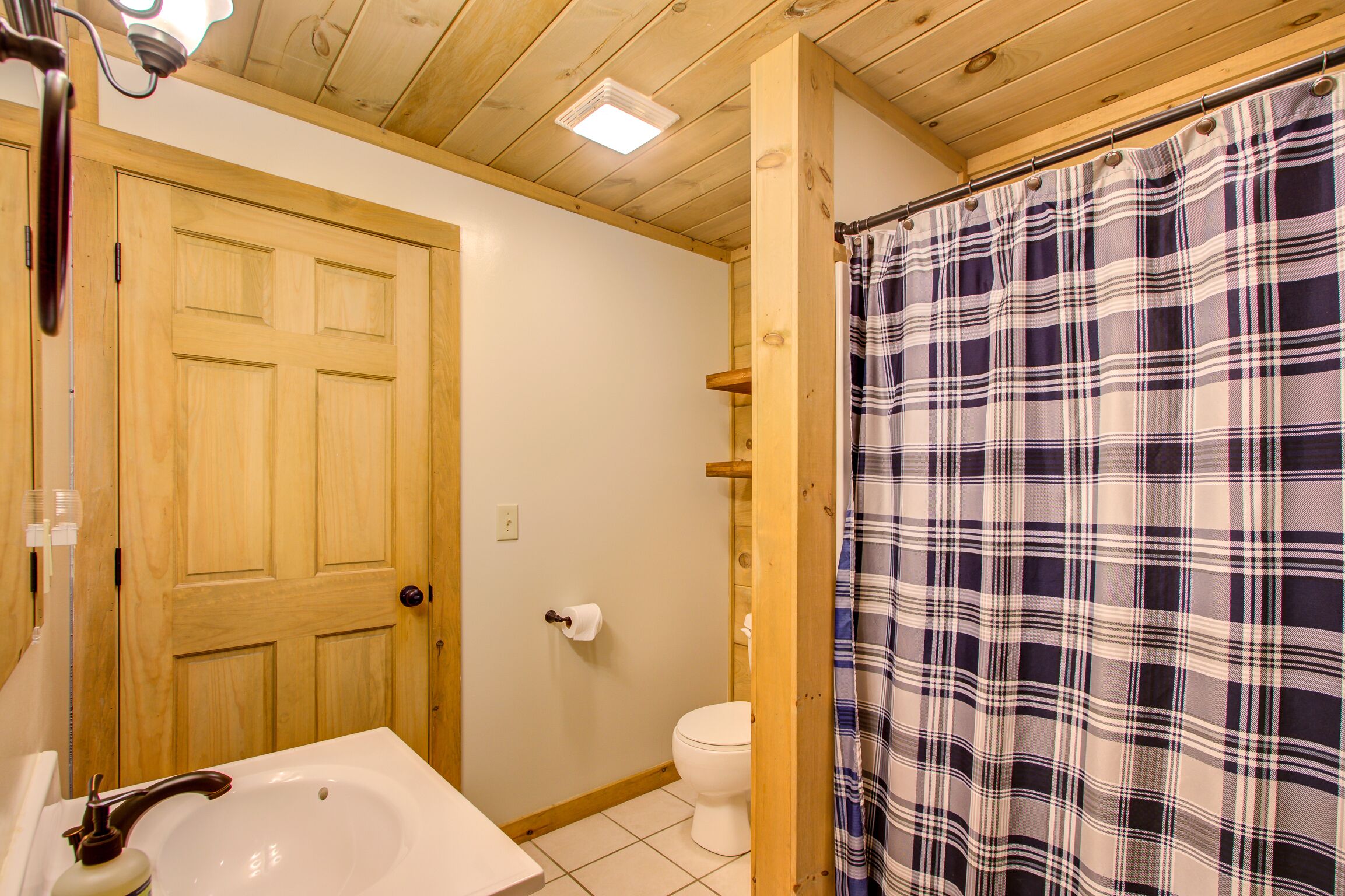 Un baño con puerta de madera, lavamanos blanco, inodoro y cortina de ducha escocesa.