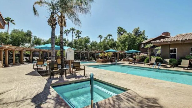 A pool area with lounge chairs and umbrellas, surrounded by palm trees and a building with a tiled roof.