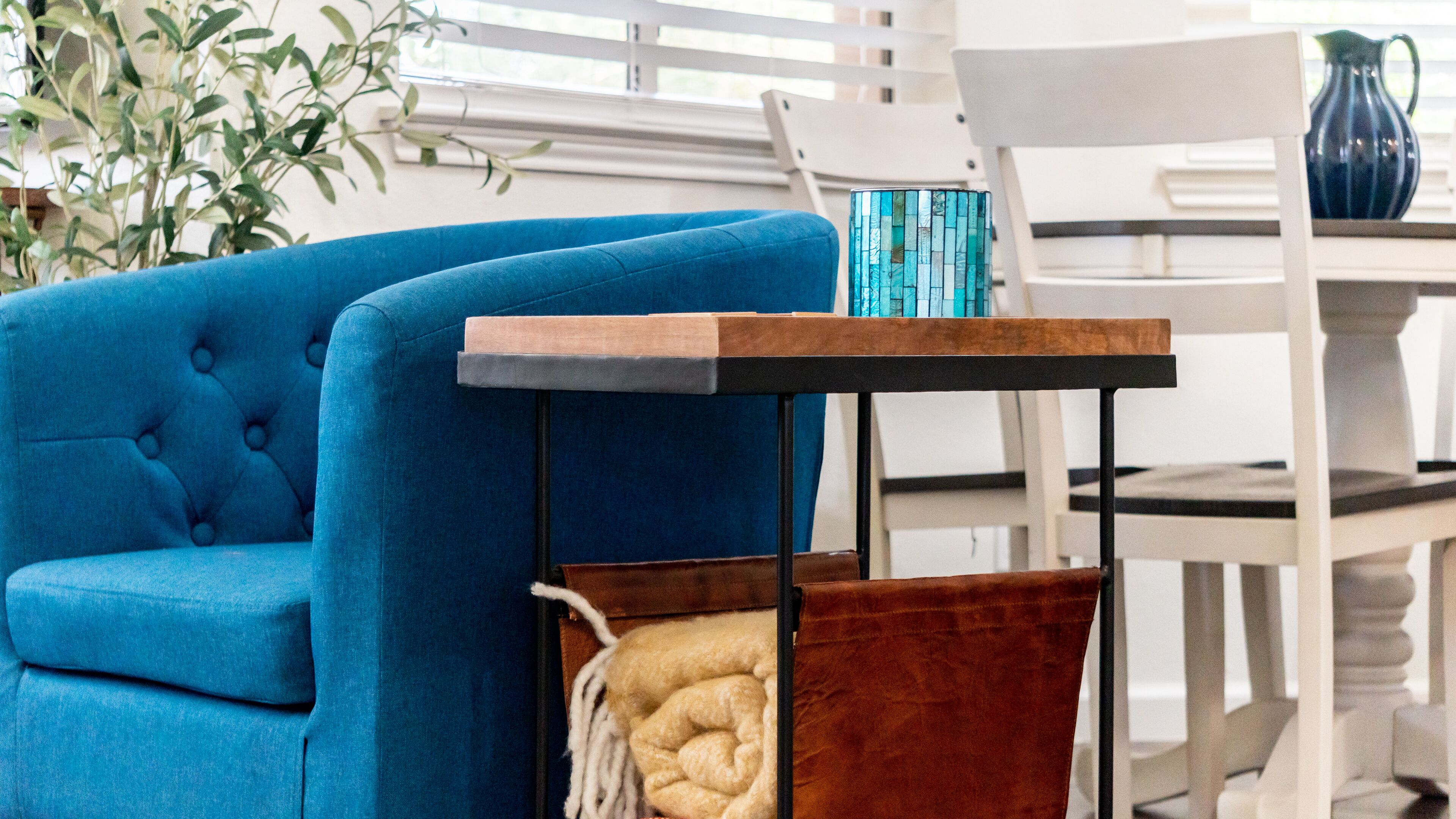 Un fauteuil bleu, une petite table d’appoint en bois et une chaise blanche se trouvent dans une pièce dotée d’une fenêtre et d’une plante.