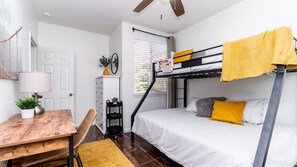 Ein kleines Zimmer mit Etagenbett, Schreibtisch mit Lampe, Stuhl, einer kleinen Pflanze und einem Fenster mit Jalousien.