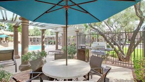 An outdoor patio with a round table and chairs, a blue umbrella, and a pool in the background.