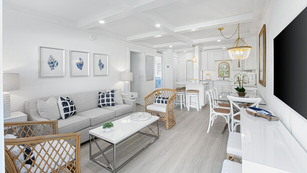 Un salon moderne avec un canapé, une table basse et des fauteuils en osier. On y trouve également un coin repas avec une table et des chaises, et une cuisine à l’arrière-plan.