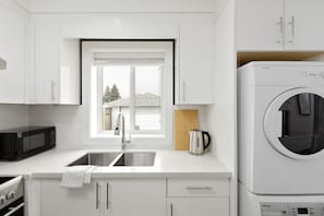 Cozinha moderna com armários brancos, uma pia de aço inoxidável, micro-ondas, chaleira e máquina de lavar de abertura frontal.