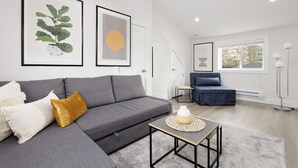 Una sala moderna con un sofá gris, una mesita de centro pequeña y un sillón azul. Hay dos obras de arte enmarcadas en la pared y una lámpara de pie.