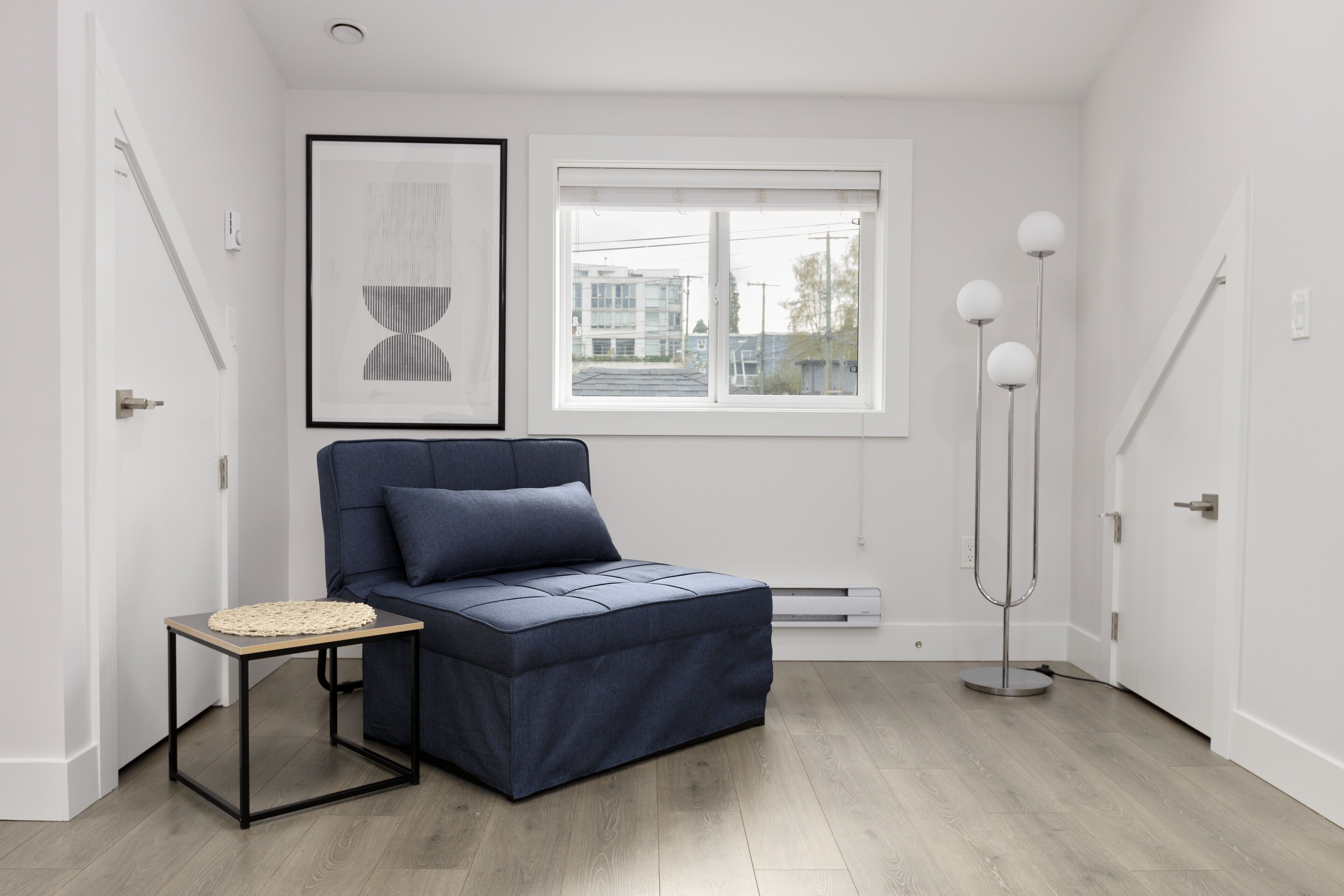 Ein modernes Zimmer mit einer Couch, einem kleinen Tisch, einer Stehlampe, einem gerahmten Kunstwerk und einem Fenster mit Blick auf Gebäude.