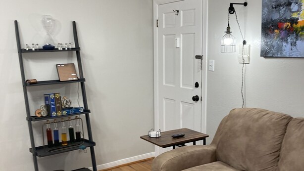 A living room with a beige sofa, a black shelving unit with books and decorative items, a white door, and a small table with a remote control. - Convenient One Bedroom Hancock Condo (Austin)