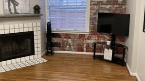A living room with a fireplace, a TV on a stand, a window with blinds, and a brick wall. - Convenient One Bedroom Hancock Condo (Austin)