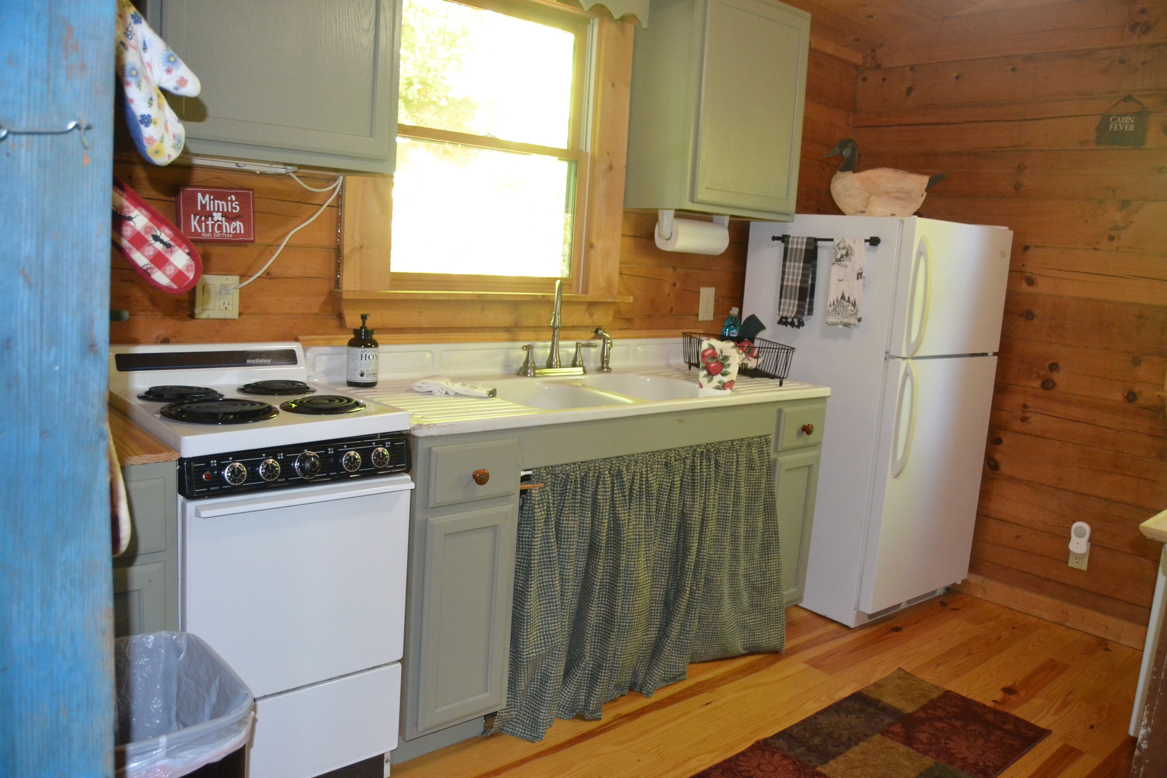 A compact kitchen with a stove, sink, and refrigerator, wooden walls, and a window.