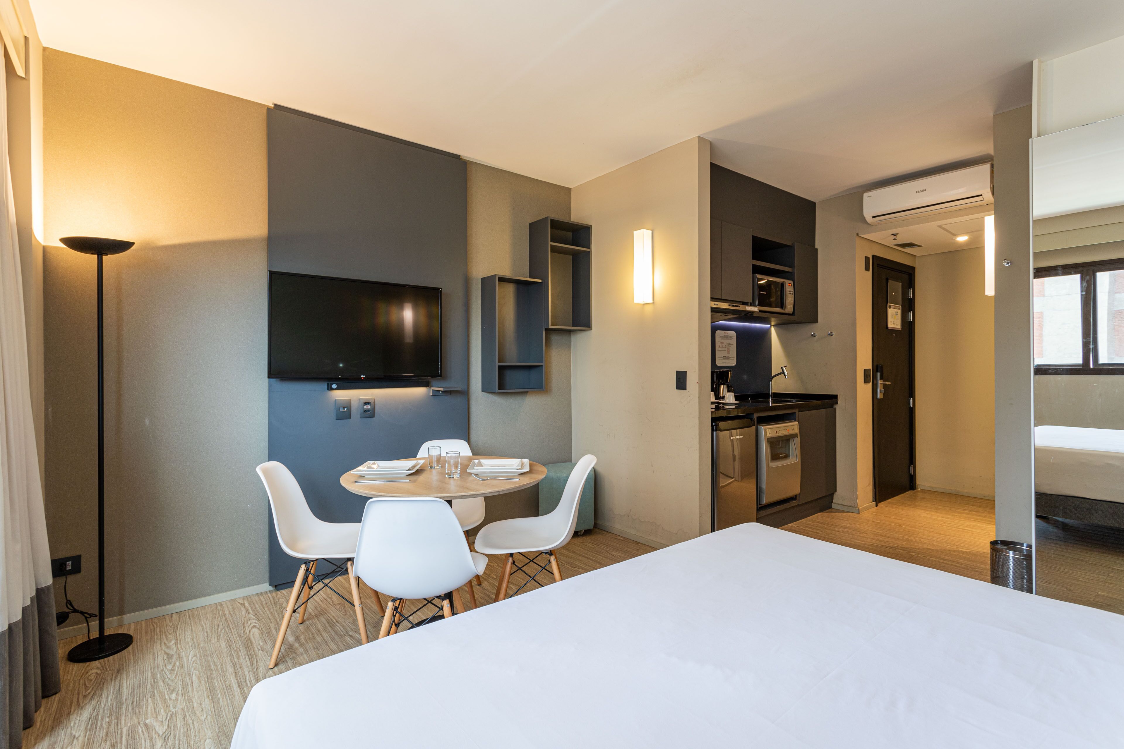 A hotel room with a bed, a dining area with chairs and a table, a TV mounted on the wall, and a kitchenette with a microwave and oven.