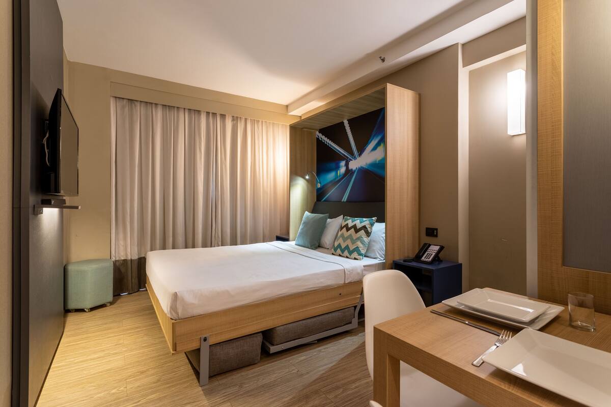 Quarto de hotel com cama, escrivaninha com cadeira, telefone e televisão na parede.