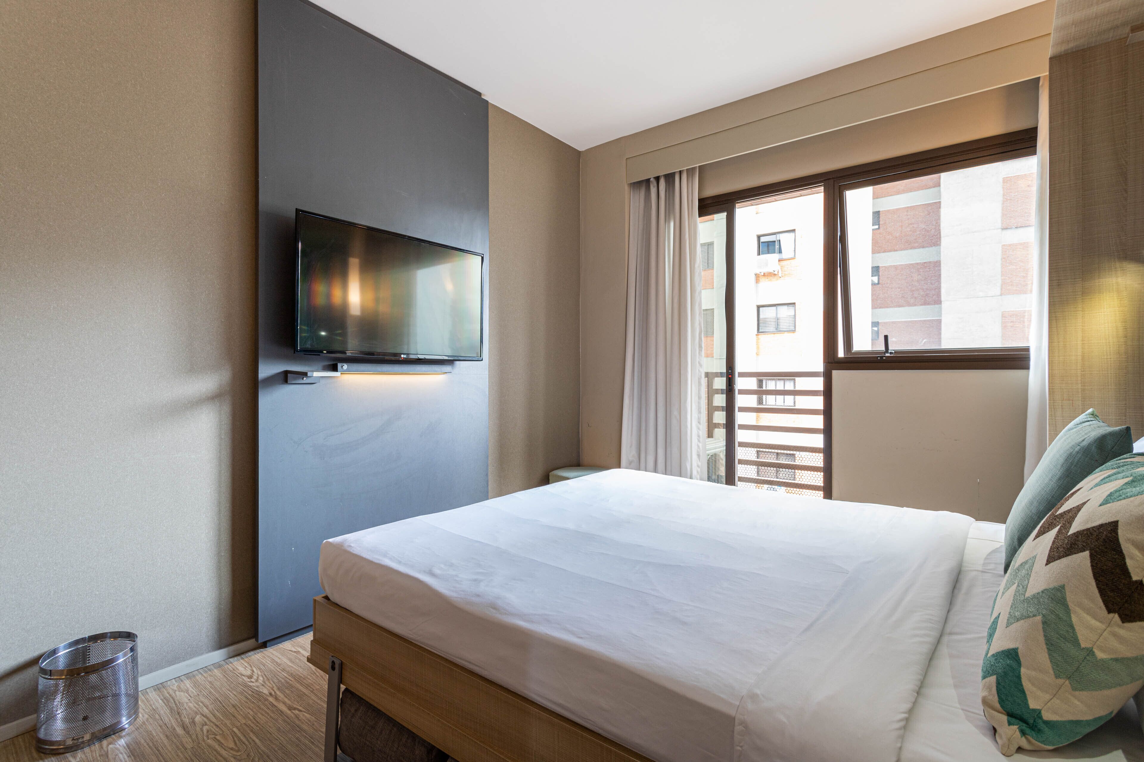 A hotel room with a bed, a TV mounted on the wall, a window with curtains, and a view of buildings outside.
