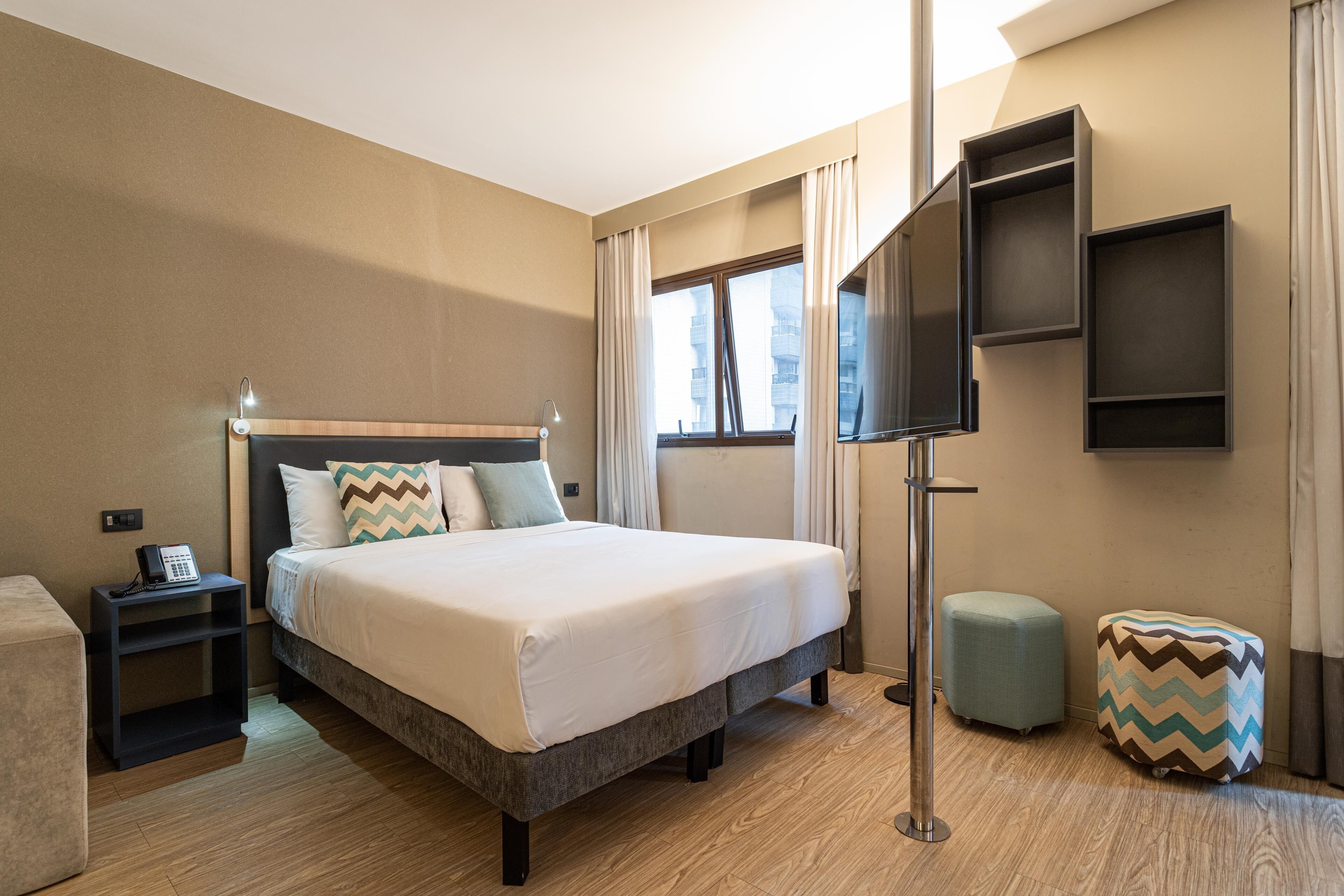 Una habitación de hotel con cama, mesita de noche, televisor y un pequeño otoman.