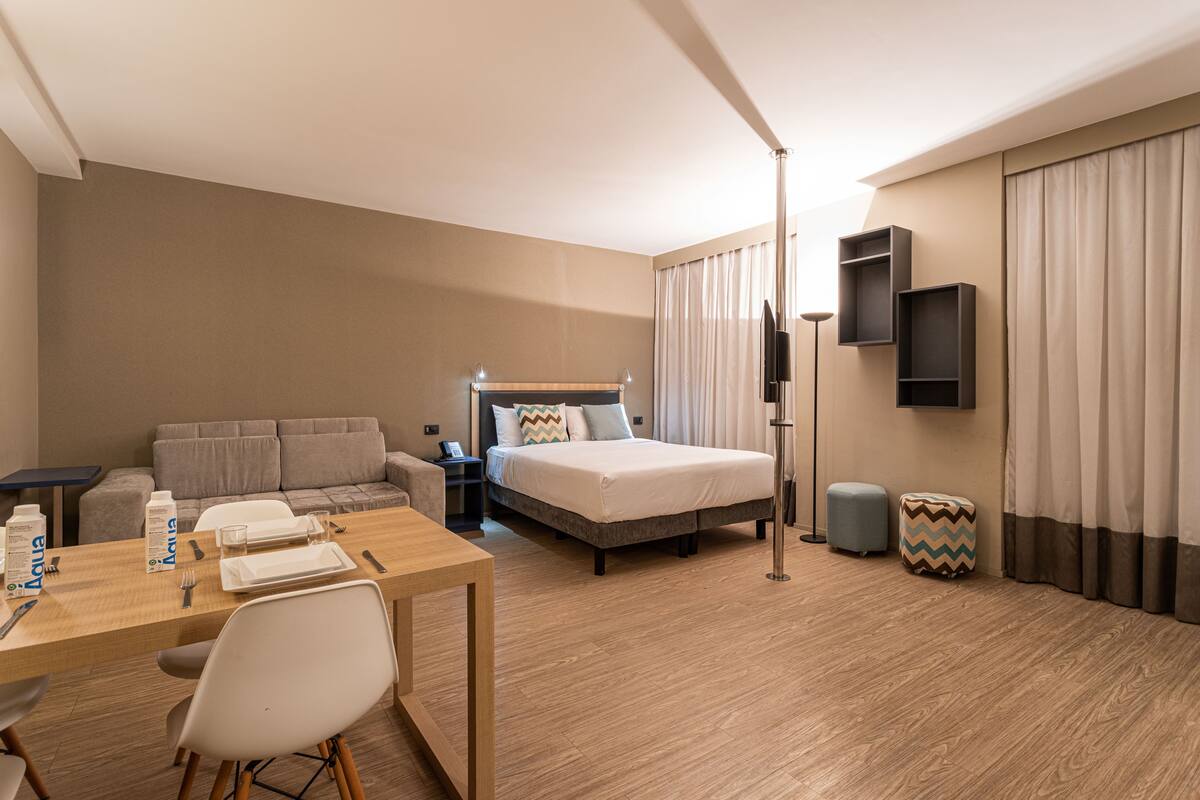 Quarto de hotel com cama, sofá, mesa de jantar com cadeiras, televisão e mesa de cabeceira.