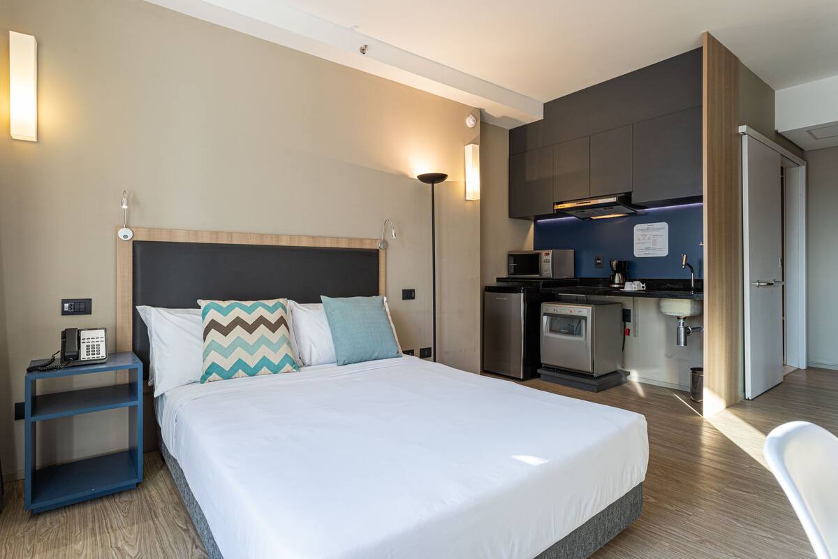 Quarto de hotel com cama, mesa de cabeceira, abajur e cozinha compacta.
