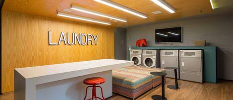 A laundry area with a wooden wall, a washing machine, a dryer, a television, and a bar stool.