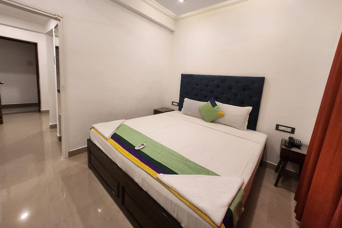 Una cama bien tendida con una colcha blanca y verde a rayas, un cabecero azul oscuro y una sola almohada verde. Hay una mesita de noche con un teléfono y una pequeña lámpara. La habitación tiene paredes blancas y un piso de baldosas.