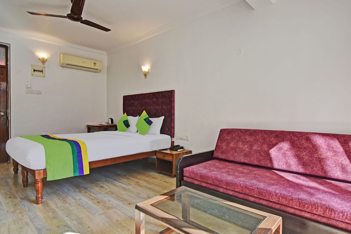 Una habitación de hotel con una cama, un sofá rojo, piso de madera, un ventilador de techo y dos aires acondicionados instalados en la pared.